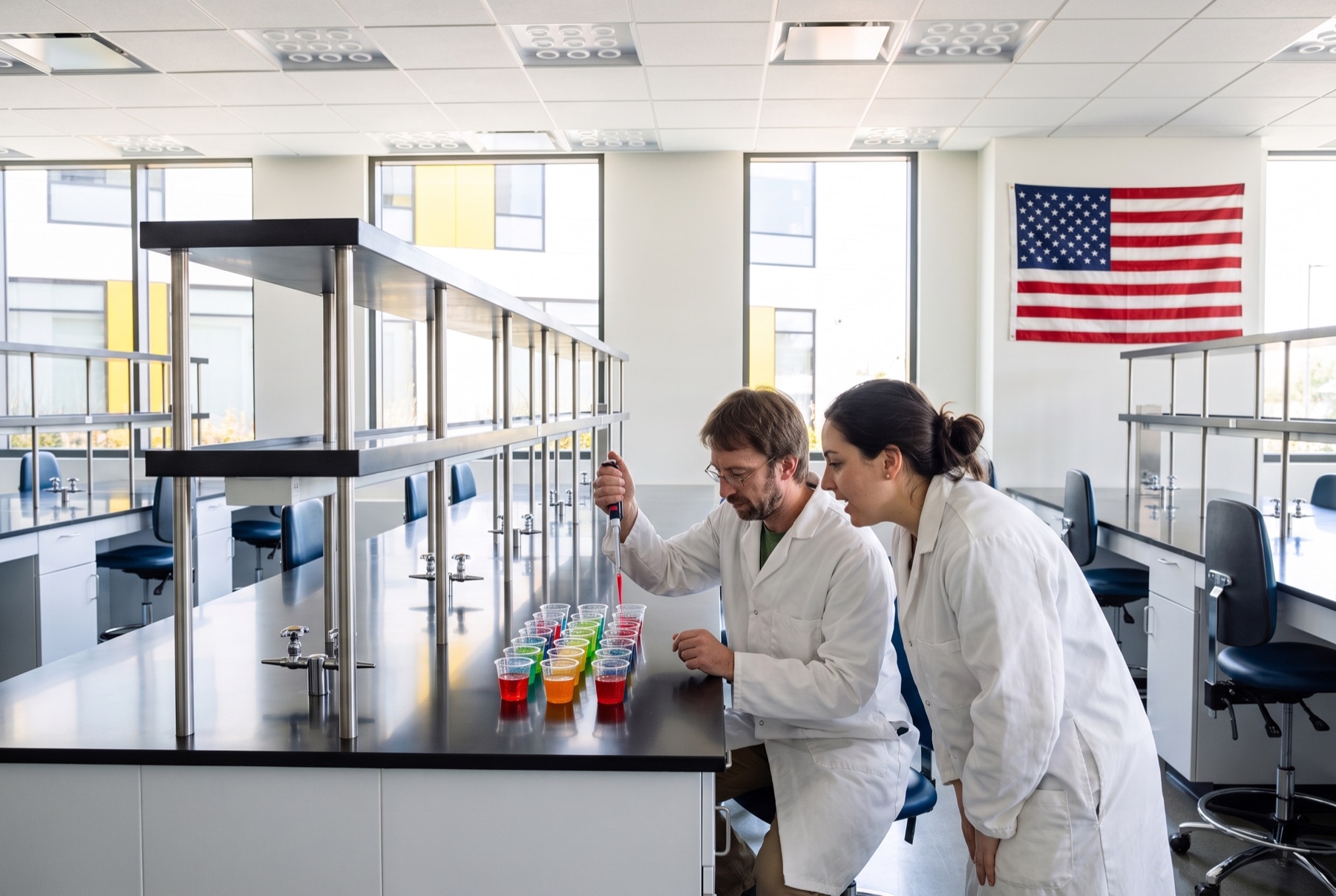 Two Tomorrow colleagues at a steel counter pipetting samples into a row of colorful tasting cups, a United States flag on the wall behind them