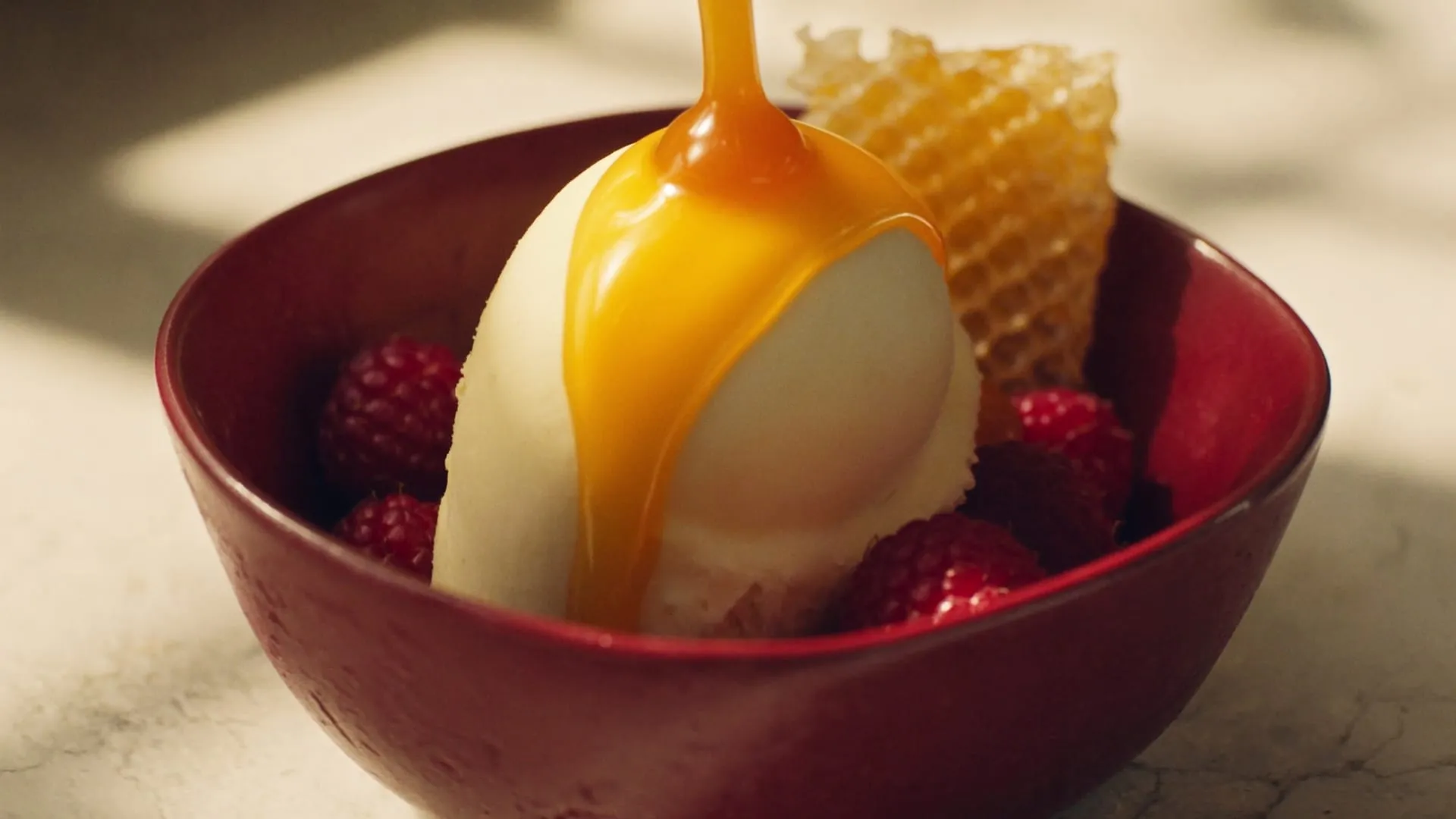 Caramel pouring over a scoop of gelato in a burgundy bowl, with raspberries and honeycomb